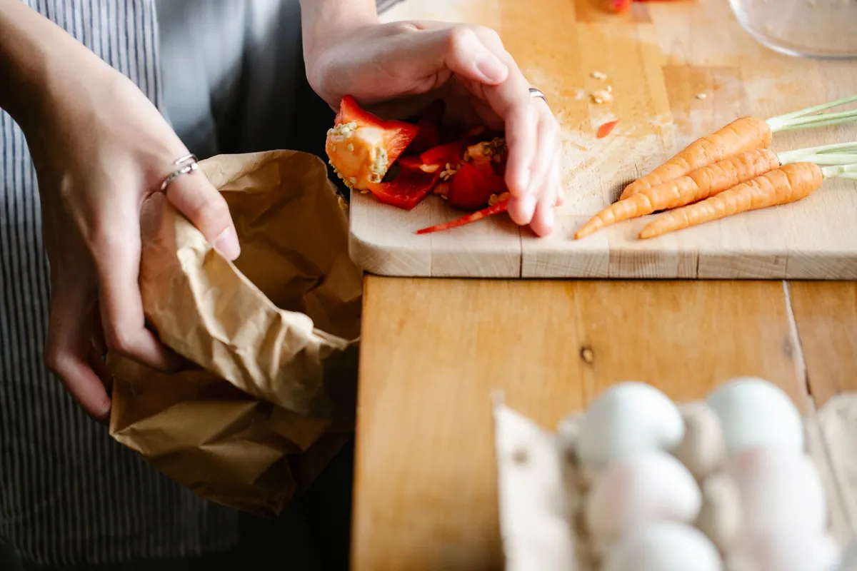 En cuisine, des mains rassemblent des épluchures et des restes de préparation dans un sac en papier