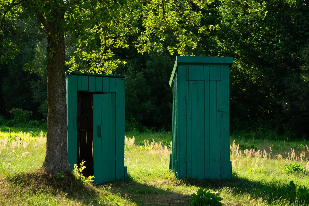 Deux cabines de toilettes sèches en bois vert installées dans une prairie, à l'ombre d'un arbre